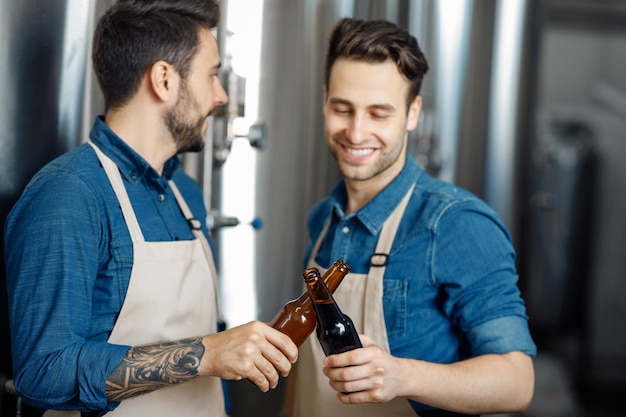 Inicio exitoso, pequeña empresa familiar y degustación de cerveza artesanal en planta. Trabajadores o propietarios alegres y atractivos del milenio en delantales, tintinean vasos de bebida, cerca de cubas de acero en el interior de la cervecería