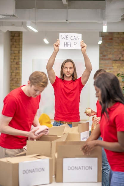 Initiative. Junger optimistischer Kerl, der Plakat in erhobenen Händen mit Inschrift und freiwilligen Kollegen zeigt, die Spendenboxen packen