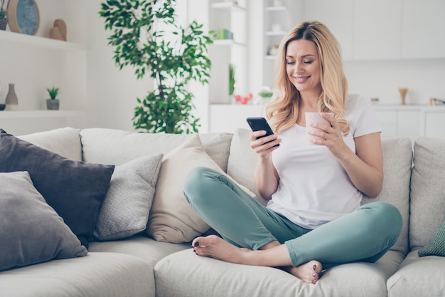 inländische hübsche fröhliche Dame, die sich entspannt auf der Couch sitzt und die Telefon hält Tasse