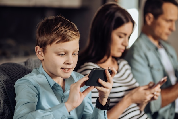 Inmerso en los juegos. La atención se centra en un agradable niño preadolescente que juega en el teléfono y sonríe con entusiasmo mientras sus padres se sientan a su lado y también usan sus teléfonos.