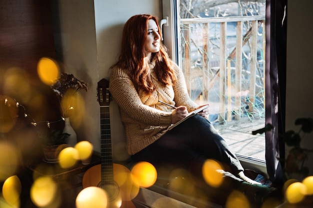 Innen gemütlich Hobbys für Winter Herbst kalte Jahreszeit Rothaarige Frau spielt Akustikgitarre und hört Musik und sitzt am Fenster ihres Hauses