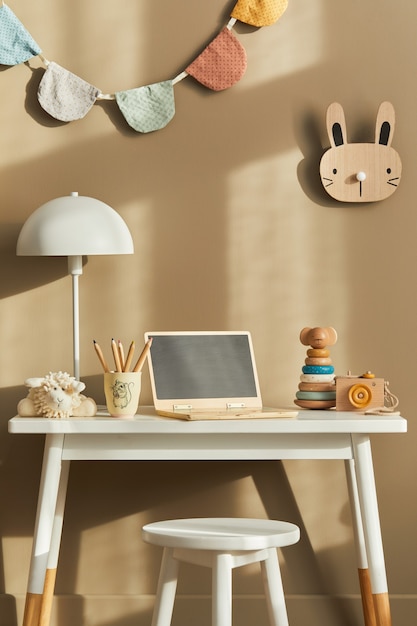 Innenarchitektur des stilvollen Kinderzimmerraums mit weißem Schreibtisch, hölzernem Laptop, Spielzeug, Kinderaccessoires, Lampe, gemütlicher Dekoration und hängenden Baumwollfahnen an der beige Wand.