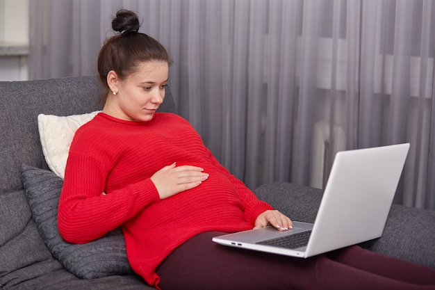 Innenaufnahme der schwangeren Frau gekleideten roten Pullover und kastanienbraune Leggings, hält Hand auf Bauch, sieht Filme online