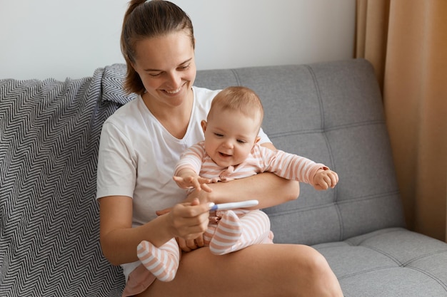 Innenaufnahme des Lächelns glücklich zufrieden posiert zu Hause im gemütlichen Wohnzimmer auf dem Sofa mit ihrem Baby in den Händen, hält positiven Schwangerschaftstest und drückt das Glück des zukünftigen Kindes aus.