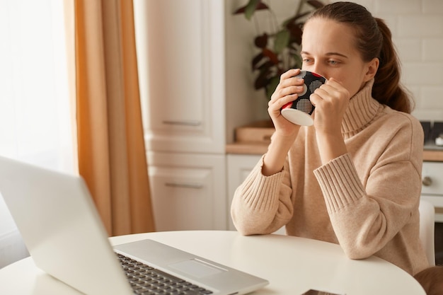 Innenaufnahme einer attraktiven Frau mit beigem Pullover, die in der Küche posiert und Kaffee oder Tee trinkt und eine Tasse in den Händen hält, die am Tisch vor einem Notizbuch sitzt