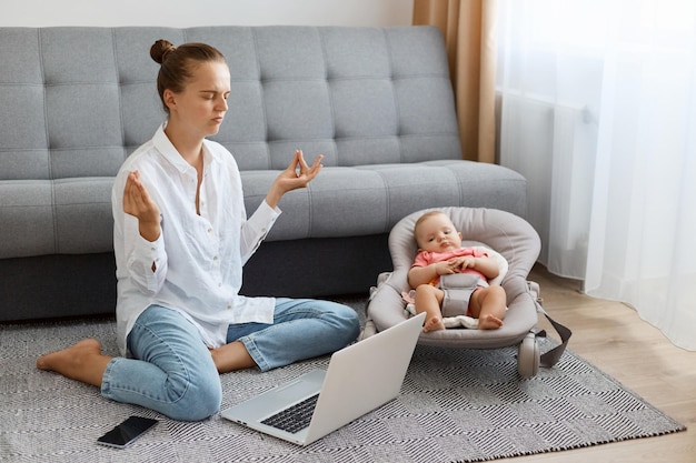 Innenaufnahme einer attraktiven, müden Frau mit Brötchenfrisur, weißem Hemd und Jeans, die vor einem Laptop auf dem Boden sitzt und sich um das Baby kümmert, das Yoga macht, um sich zu beruhigen