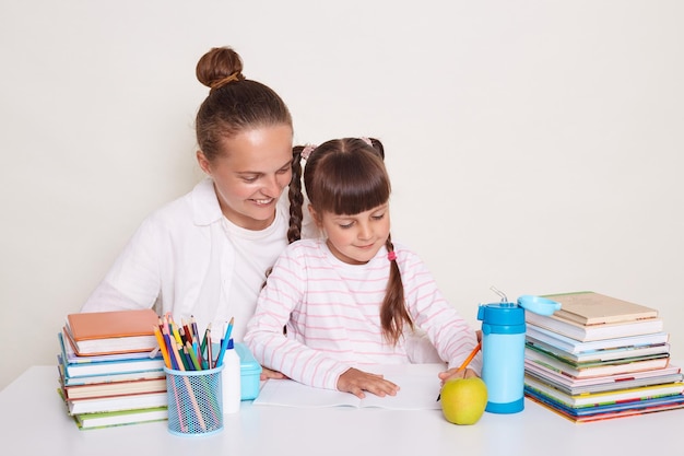 Innenaufnahme einer attraktiven Mutter mit ihrer Tochter, die am Tisch mit Büchern sitzt, die Hausaufgaben machen