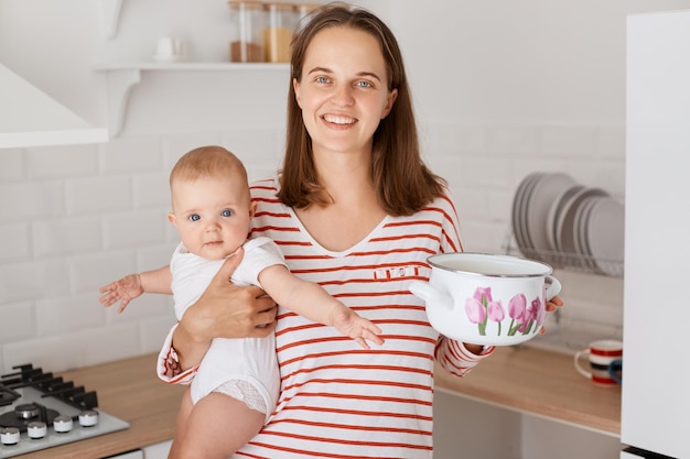 Innenaufnahme einer dunkelhaarigen Frau und eines kleinen Kindes in der Küche zu Hause, Frau in gestreiftem Hemd, die Baby in den Händen hält und mit ihrem Kind im Topf kocht, um Glück auszudrücken