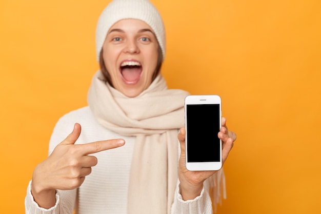 Innenaufnahme einer erstaunten, aufgeregten Frau, die in Mütze steht und in einen Schal gehüllt ist, isoliert vor gelbem Hintergrund, der ein Handy mit leerem Display zeigt, Werbefläche, Kopierfläche für Werbung