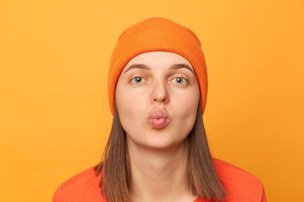 Innenaufnahme einer flirtenden romantischen Hipster-Frau mit orangefarbenem Pullover und Hut, die Luftküsse sendet und in die Kamera blickt, die isoliert vor gelbem Hintergrund posiert