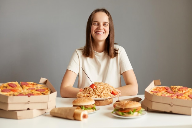 Innenaufnahme einer Frau mit braunen Haaren in weißem T-Shirt, die isoliert vor grauem Hintergrund am Tisch sitzt und von leckerem Fast Food umgeben ist, das in die Kamera lächelt