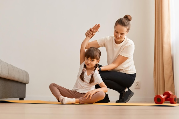 Innenaufnahme einer Frau mit kleinem Mädchen in Sportkleidung, die zu Hause Sportübungen macht Kind sitzt auf Matte in Yoga-Pose und dehnt ihren Körper, um die Flexibilität zu verbessern Mutter hilft ihrer Tochter