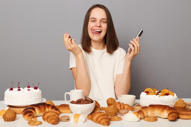 Innenaufnahme einer fröhlichen, attraktiven Frau mit braunen Haaren, die ein weißes T-Shirt trägt und am Tisch sitzt, mit Telefon und Keks in der Hand, die die Zunge herausstreckt und hungrig ist, isoliert auf grauem Hintergrund