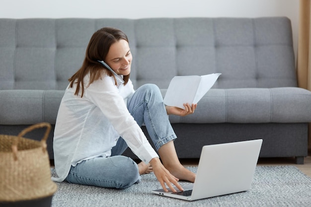 Innenaufnahme einer fröhlichen Frau mit weißem Hemd und Jeans, die mit einem Laptop und einem Telefon auf dem Boden neben einem Sofa sitzt und ihre Freude bei der Arbeit und einem angenehmen Gespräch ausdrückt