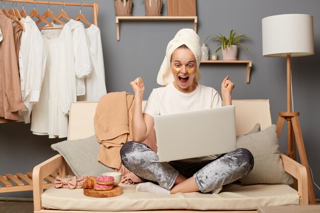 Innenaufnahme einer fröhlichen, überglücklichen Frau mit weißem T-Shirt, grauer Hose und einem Handtuch auf dem Kopf, die auf Husten sitzt, mit einem Notizbuch, das auf den Bildschirm schaut, und geballten Fäusten, die sich über das Wetten und Gewinnen freuen