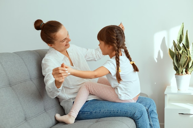 Innenaufnahme einer glücklich lächelnden Frau mit weißem Hemd und Jeans, die mit ihrer charmanten Tochter auf dem Sofa sitzt, die Familie zusammen spielt und sich gegenseitig die Hände hält.