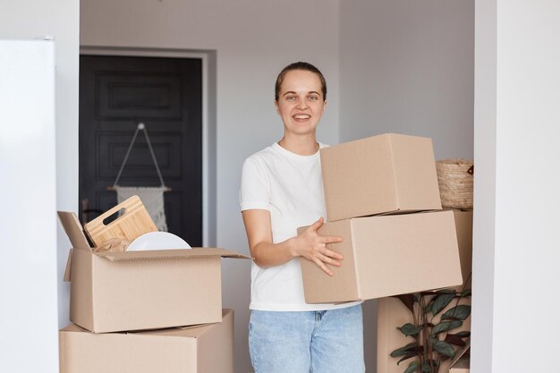 Innenaufnahme einer glücklich lächelnden Frau mit weißem T-Shirt und Jeans im lässigen Stil, die mit Kartons mit Habseligkeiten steht, mit positivem Ausdruck in die Kamera schaut und froh ist, umzuziehen.