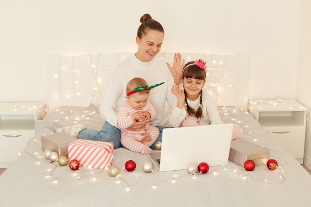 Innenaufnahme einer glücklichen attraktiven Frau und zweier Kinder, die Laptop-Computer verwenden, um per Videoanruf zu sprechen, während sie im Bett mit Weihnachtsdekoration sitzen und die Hand zur Notebook-Webkamera winken.