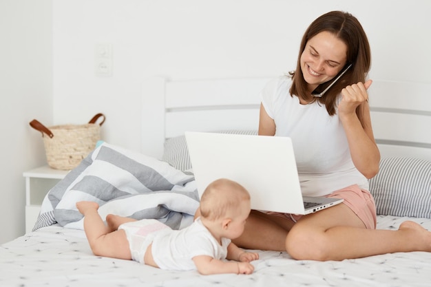 Innenaufnahme einer glücklichen, fröhlichen Frau, die mit ihrer kleinen Tochter auf dem Bett sitzt und online arbeitet und einen Laptop und ein sprechendes Handy mit geballter Faust hält, um den Erfolg zu feiern