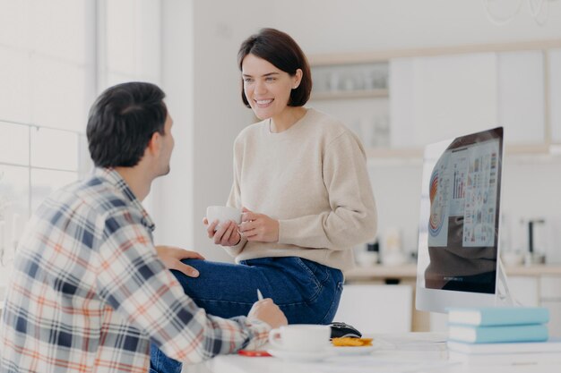 Innenaufnahme einer glücklichen jungen Frau und eines Mannes, die etwas mit einem Getränk besprechen. Verwenden Sie einen modernen Computer, um Finanzberichte in einem geräumigen hellen Raum zu erstellen