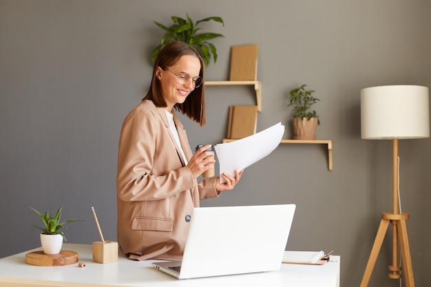Innenaufnahme einer hübschen, süßen, braunhaarigen Frau mit beigefarbener Jacke, die im Büro posiert und Informationen in Dokumenten liest, indem sie mit dem Handy Kaffee trinkt