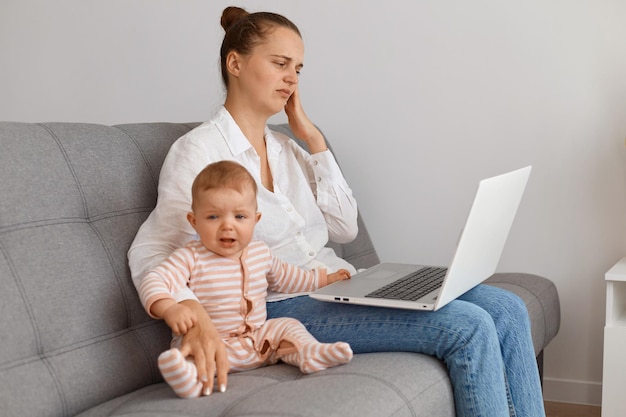 Innenaufnahme einer jungen erwachsenen dunkelhaarigen Frau mit Laptop auf dem Sofa mit ihrem Baby, die Fernarbeit macht, Stress verspürt, müde ist zu arbeiten und sich um ihr Kind kümmert.