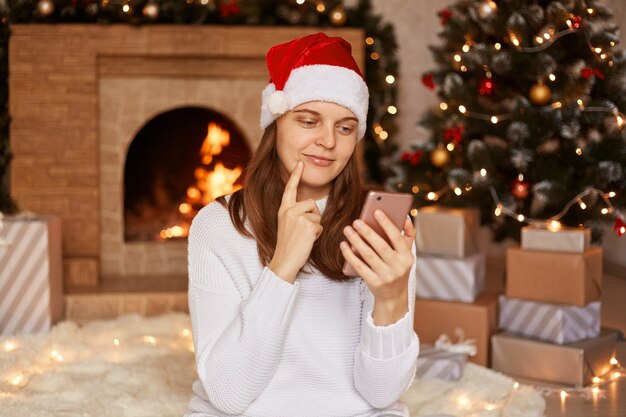 Innenaufnahme einer jungen erwachsenen Frau mit weißem Pullover und Weihnachtsmann-Hut, die in einem festlichen Raum in der Nähe von Weihnachtsbaum und Kamin sitzt, das Telefon benutzt und an das neue Jahr denkt und gratuliert.