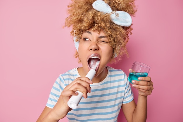 Innenaufnahme einer jungen Frau mit lockigen Haaren, die Zähne mit einer elektrischen Bürste putzt, hält ein Glas Mundwasser, zwinkert dem Auge zu, das in einem lässig gestreiften T-Shirt gekleidet ist, das über rosafarbenem Hintergrund isoliert ist Mundhygienekonzept