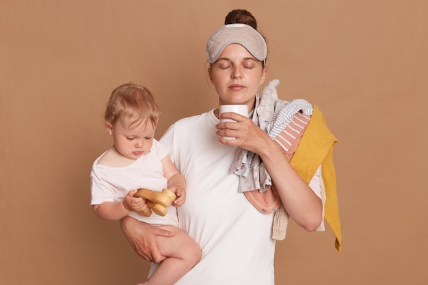 Innenaufnahme einer jungen, müden Mutter und eines kleinen Kindes nach einer schlaflosen Nacht Erschöpfte Frau mit Baby, die mit Kaffee steht, isoliert über braunem Hintergrund Postpartale Depression im Mutterschaftsurlaub