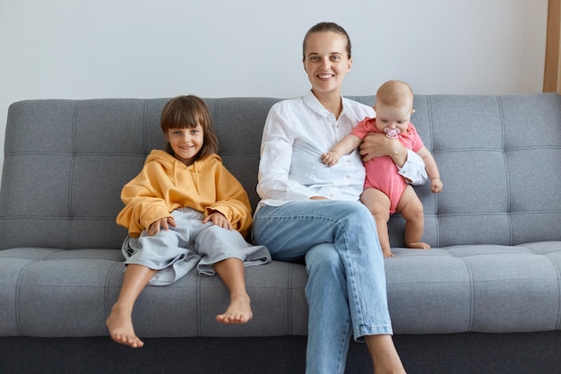 Innenaufnahme einer jungen Mutter, die ein weißes Hemd und Jeans trägt, während ihre Kinder auf dem Sofa sitzen und mit glücklichem, positivem Gesichtsausdruck in die Kamera schauen. Familie verbringt Zeit miteinander