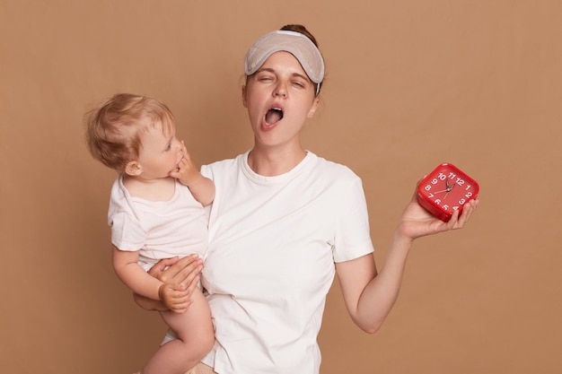 Innenaufnahme einer jungen Mutter mit Brötchenfrisur, die ihre kleine Tochter und einen roten Wecker hält, die schläfrig gähnend mit geschlossenen Augen und mit Augenbinde isoliert vor braunem Hintergrund steht