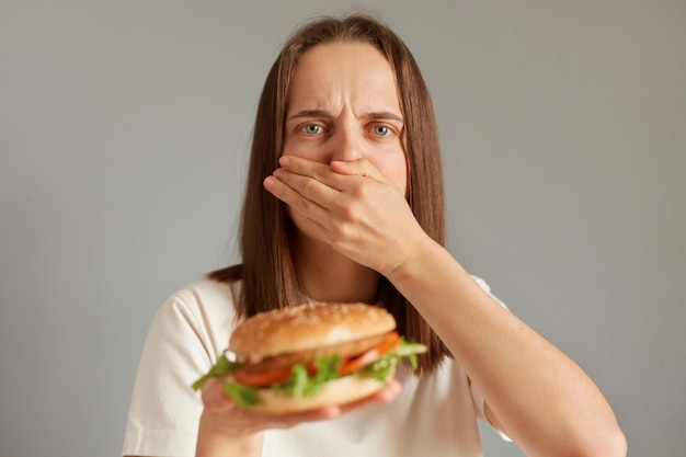 Innenaufnahme einer kranken Frau, die sich krank fühlt, wenn sie Fast Food sieht, das ihren Mund bedeckt, und Übelkeit verspürt, wenn sie verdorbenes Sandwich isst und in die Kamera schaut und sich unwohl fühlt