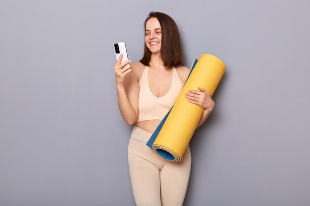 Innenaufnahme einer lächelnden, begeisterten, sportlichen Frau, die Sportkleidung mit Fitnessmatte in den Händen trägt, isoliert auf grauem Hintergrund, indem sie mit ihrem Smartphone die verbrannten Kalorien in der App überprüft