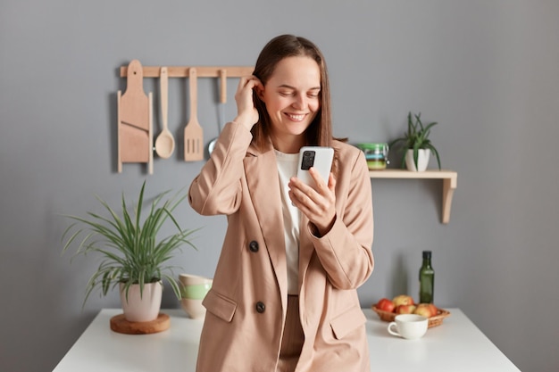 Innenaufnahme einer lächelnden, erfreuten, braunhaarigen Frau im beigen Anzug, die zu Hause neben dem Tisch in der Küche steht und mit dem Handy soziale Netzwerke beim Surfen im Internet überprüft