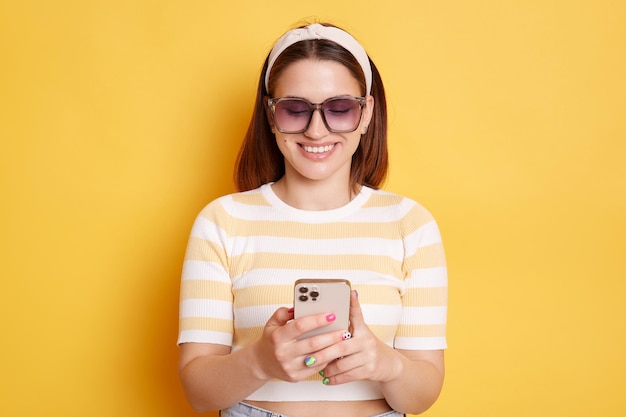 Innenaufnahme einer lächelnden, erfreuten Frau mit Sonnenbrille, gestreiftem T-Shirt und Haarband, die isoliert vor gelbem Hintergrund posiert und das Smartphone hält, das im Internet surft und soziale Netzwerke überprüft