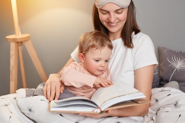 Innenaufnahme einer lächelnden europäischen Frau mit weißem T-Shirt und Augenbinde, die im Bett sitzt, während ihre kleine Tochter Mama Märchen für Kinder liest, die ein Buch in den Händen halten