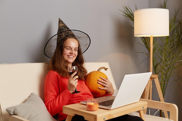 Innenaufnahme einer lächelnden, festlichen Frau mit Pullover und Hexenhut, die auf dem Sofa sitzt und mit einem Laptop Wein aus Glas trinkt, um Halloween mit ihren Freunden per Videoanruf zu feiern