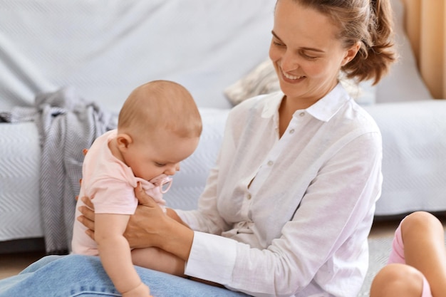 Innenaufnahme einer lächelnden glücklichen Frau mit dunklem Haar, die zu Hause mit ihrer kleinen Tochter spielt, ein weißes Hemd trägt, mit einem süßen Baby in den Händen sitzt und ihr Kind ansieht.