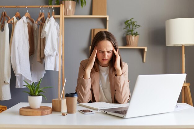 Innenaufnahme einer müden, erschöpften Designerin in Jacke, die im Home Office an einem Notebook arbeitet, gegen Kleider, die im Regal hängen und unter schrecklichen Kopfschmerzen leiden, fühlt starke Schmerzen, wenn sie lange Stunden arbeitet