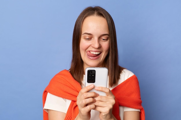 Innenaufnahme einer positiven Frau, die ein weißes T-Shirt und einen orangefarbenen Pullover trägt, der über die Schultern gebunden ist und isoliert auf blauem Hintergrund steht, mit einem Handy, das glücklich lächelt und die Zunge herausstreckt