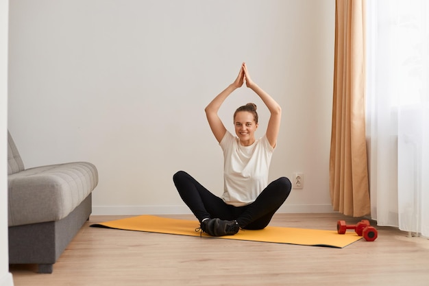 Innenaufnahme einer ruhigen Frau, die in Lotus-Pose auf einer Matte sitzt, ein weißes T-Shirt und schwarze Leggins trägt, die Arme erhebt, die Handflächen zusammenhält und mit einem charmanten, glücklichen Lächeln in die Kamera blickt.