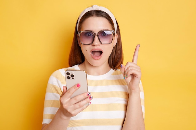 Innenaufnahme einer schockierten, erstaunten Frau mit gestreiftem Hemd, die isoliert vor gelbem Hintergrund posiert, mit einem Handy, das ein Smartphone hält und den erhobenen Finger nach oben hält, um den Mund offen zu halten