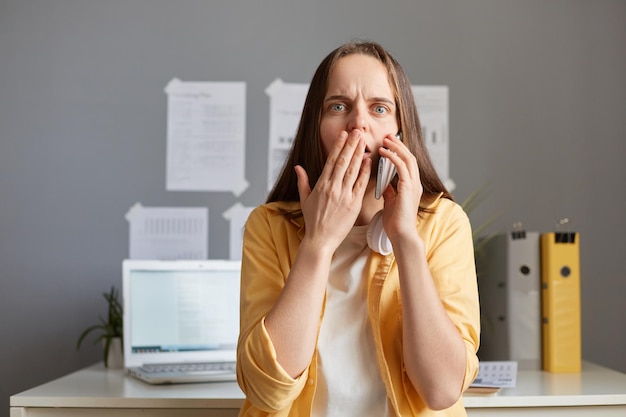 Innenaufnahme einer schockierten, erstaunten kaukasischen Frau mit braunen Haaren, die im Büro an ihrem Arbeitsplatz sitzt und mit einem gelben Hemd telefoniert und überraschte Nachrichten hört, die den Mund mit der Handfläche bedecken