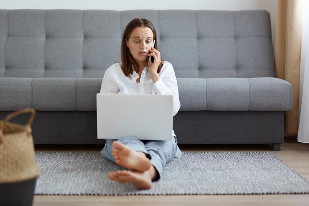 Innenaufnahme einer schockierten, überraschten, attraktiven Frau in Jeans und weißem Hemd, die an ihrem Laptop arbeitet oder per Smartphone studiert und spricht, um ihr Erstaunen auszudrücken