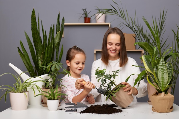 Innenaufnahme einer schönen Floristin in weißem Hemd mit braunen Haaren, die Zeit mit ihrer Tochter im Gewächshaus verbringt Mutter und kleines Kind, die Blumen neu pflanzen, um positive Emotionen auszudrücken