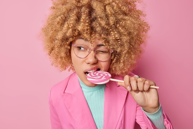 Innenaufnahme einer schönen Frau, die Naschkatzen beißt, köstliche Süßigkeiten auf einem Stock zwinkert, das Auge genießt, leckeren Lutscher zu essen, trägt eine transparente Brille und formelle Kleidung, isoliert über rosa Hintergrund