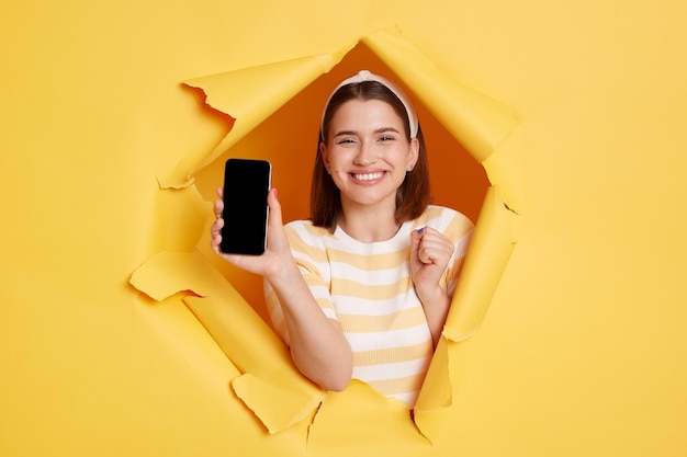 Innenaufnahme einer süßen, lächelnden, zufriedenen Frau mit gestreiftem T-Shirt, die durch ein Loch in einer gelben Papierwand blickt, die ein Smartphone mit leerem Display zeigt, Kopienraum für Werbung