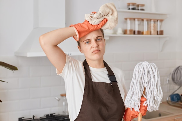 Foto innenaufnahme einer traurigen, müden frau mit dunklem haar, die ein weißes t-shirt und eine braune schürze trägt und einen mopp und ein tuch in den händen hält