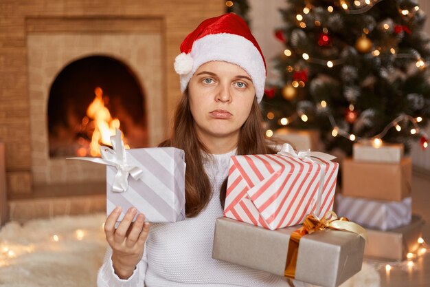 Innenaufnahme einer traurigen unglücklichen Frau mit weißem Pullover und Weihnachtsmann-Hut, die sich in einem festlichen Raum mit Kamin und Weihnachtsbaum befindet, den Geschenkkartons der Kamera zeigt und sich über Geschenke verärgert.