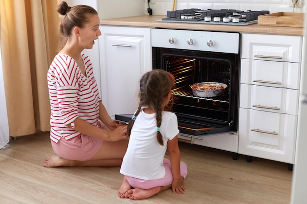 Innenaufnahme einer überraschten, erstaunten Frau mit gestreiftem Hemd und einem Kind im weißen T-Shirt, das auf dem Boden in der Nähe des Ofens sitzt und mit großen Augen nach innen schaut, die Muffins oder Kuchen backen